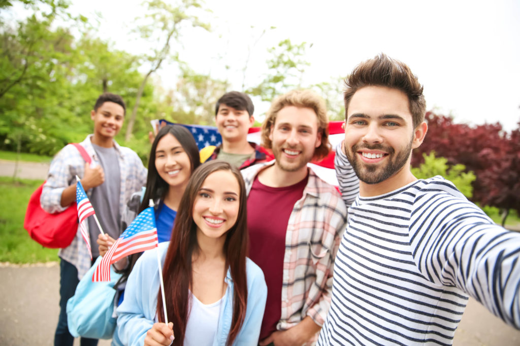 A large group of male and female college students holding small and large USA flags taking selfie outdoors. They're enjoying an educational travel experience and study abroad program with Students Fare.
