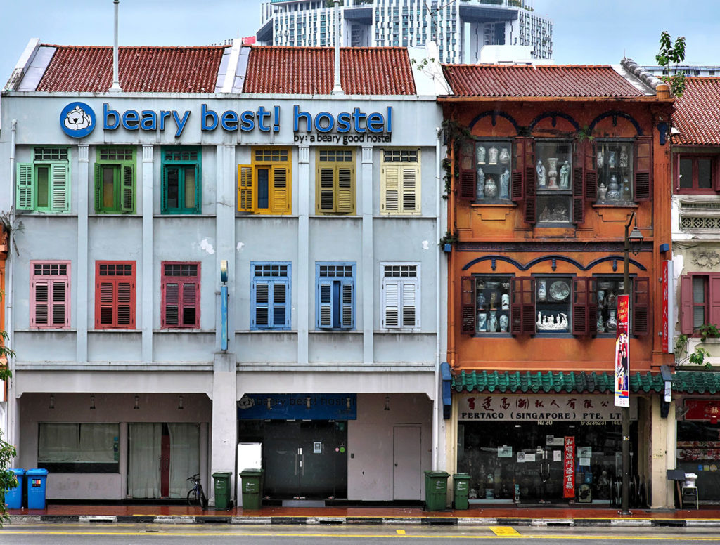 The featured image for the travel blog, Student Fare, article "hostel 101 guide." The image is of a three story hostel in Singapore next to the sidewalk where student travelers pass by.