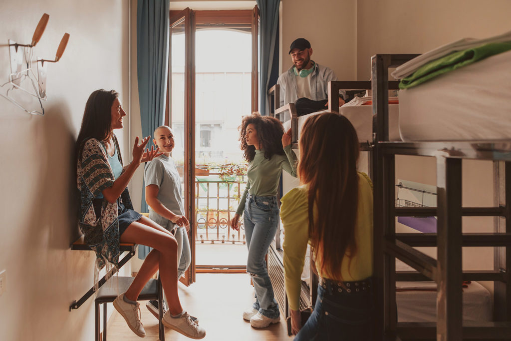A group of young student travelers gather in their hostel and have fun meeting new people. There are bunk beds in the room for student accommodations and a small bench that a young woman is sitting on.