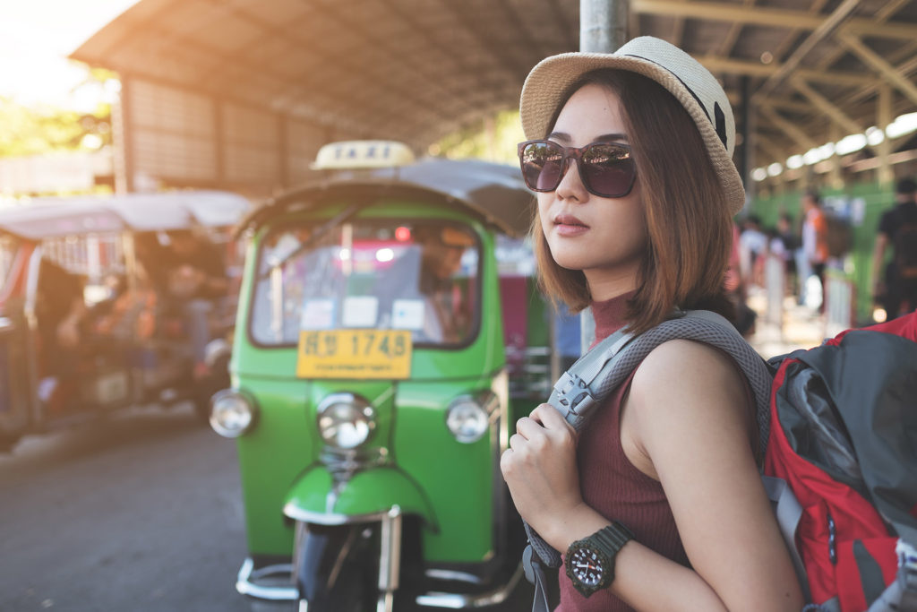 A young female traveler on an educational tour for university stands at the bus station, looking out for her transportation. In the background, a green taxi is waiting to pick up the student traveler and bring them to their hostel.