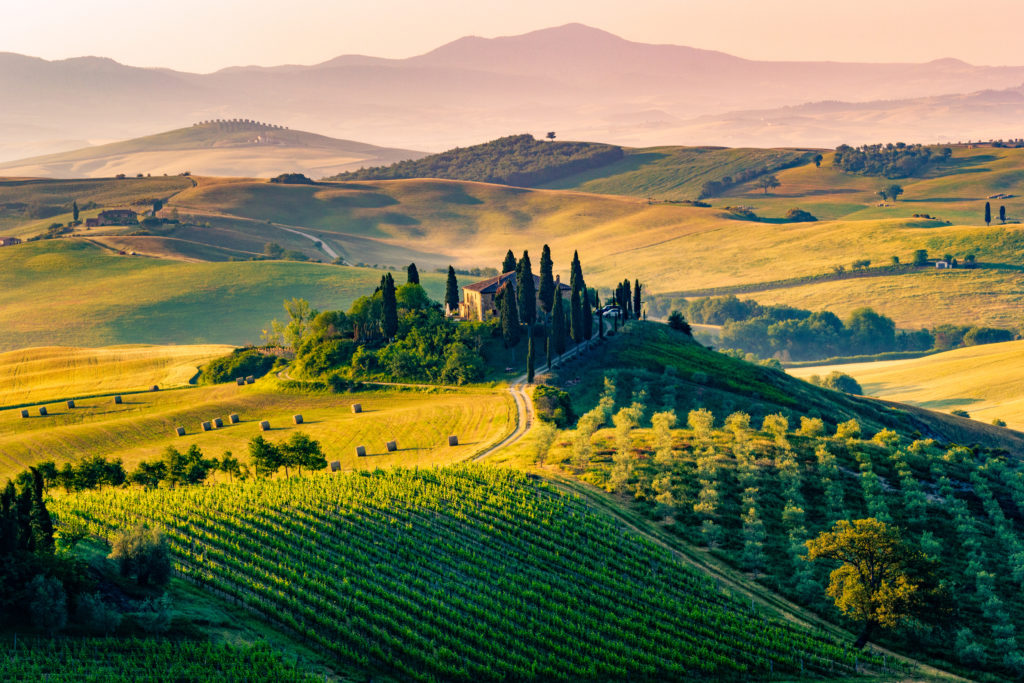 A beautiful image of the landscape in Tuscany, Italy with rolling green fields highlighted by the sunset and a small Italian villa on a hill. This image is featured in the Students Fare article about student travel called top fall break destinations.