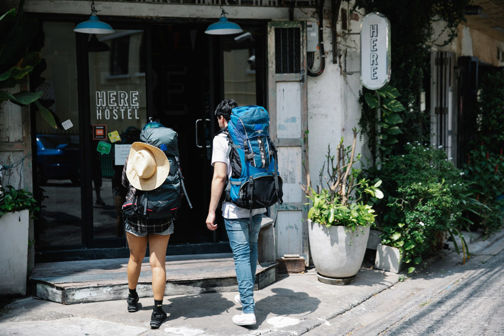 Two young students with hiking backpacks walk towards the entrance of a hostel for cheap accommodations. This is the featured image of the article "hostel 101 guide" from the Students Fare travel blog.