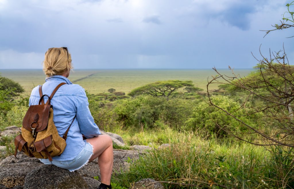 A young blonde woman sits on a rock with her back to the camera as she looks out at the African grasslands on her introverted solo adventure. This image is featured in the Students Fare student travel blog, "Best Destinations for Solo Travelers," which lists places for individual tours.