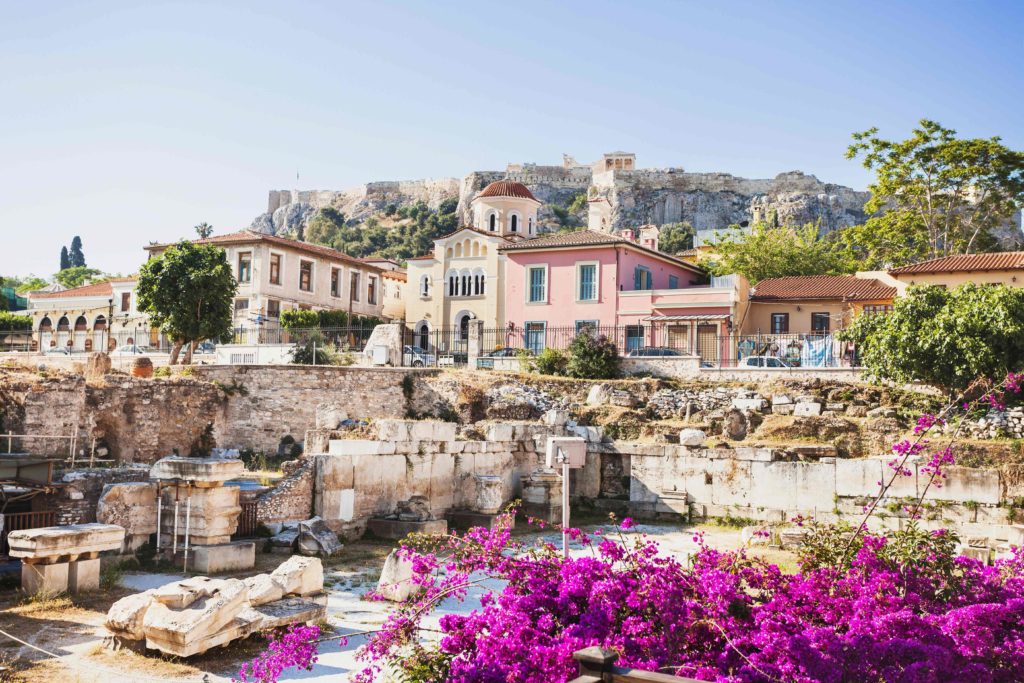 A beautiful wide shot of a small costal town Athens, Greece with pastel houses, rock cliffs, ancient ruins, and purple wildflowers. This image is used in the Students Fare student travel blog to list the top spring break destinations for college students in 2023!
