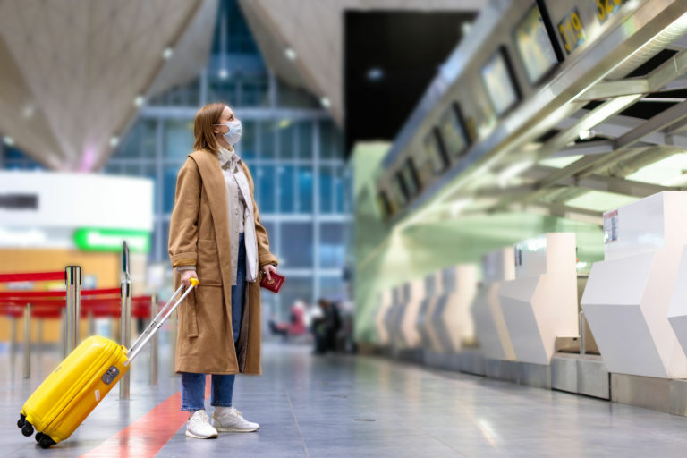 An image of a young female traveler wearing a mask and carrying a yellow suitcase, she is pausing to look at the arrival/departure board at the airport. This image is used in the Students Fare student travel blog to share with first-time flyers, a guide to international flights for students!