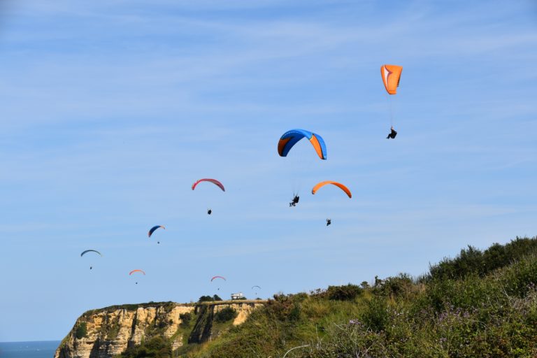 On a clear and sunny day, a large group paragliding tour flies over a seaside cliff enjoying the fun activity. This image is used in the Students Fare student travel blog, "Best Paragliding Destinations," which is a travel guide for paragliding all around the world.