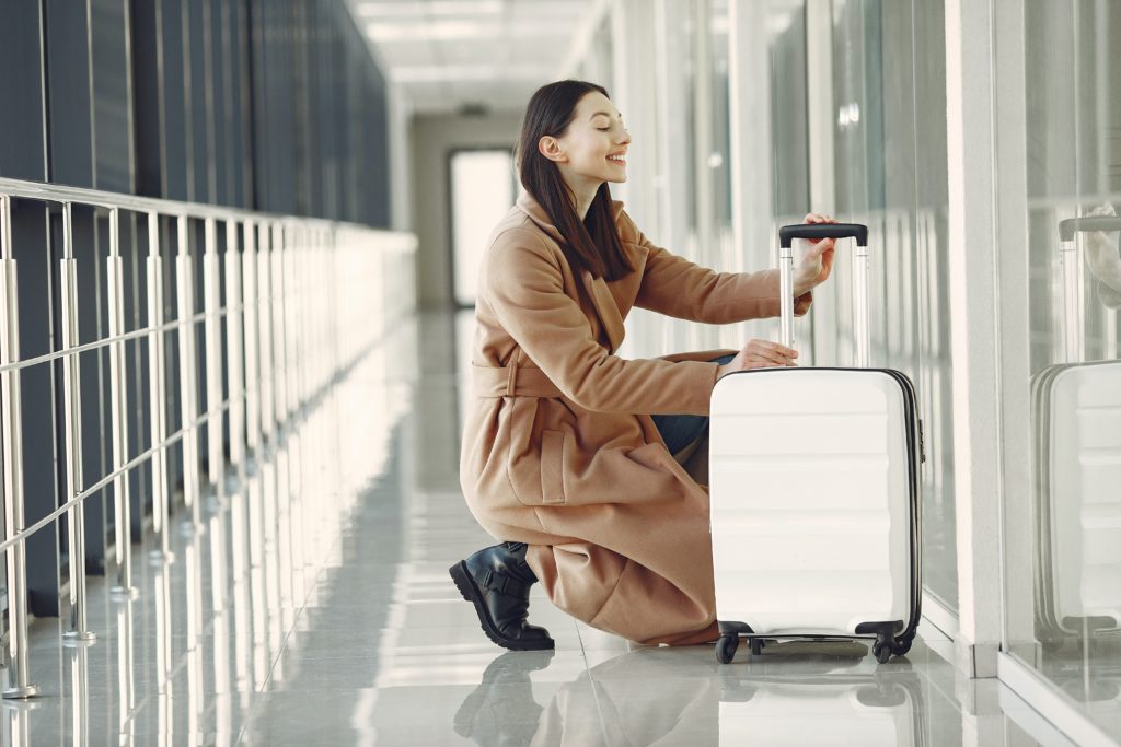 An image of a young female traveler in a brown coat with black boots kneels down to examine her white suitcase in an airport hallway. This image is used in the Students Fare student travel blog to share with first-time flyers, a guide to international flights for students!
