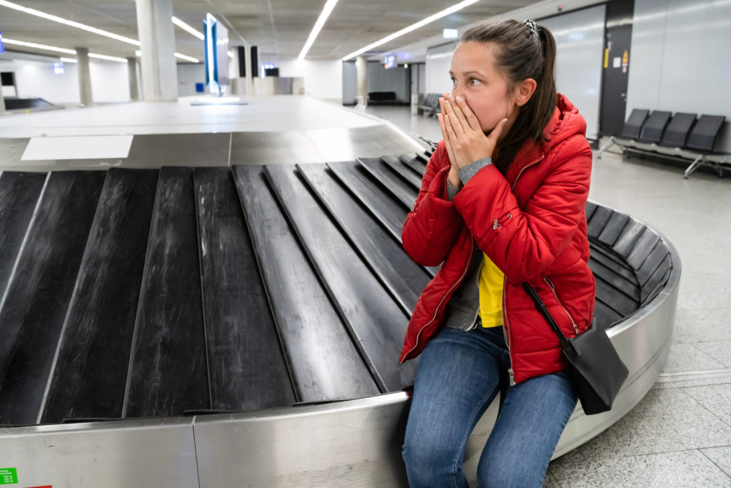 A young student traveler in the airport is stressed and upset at baggage claim because her baggage was lost and destroyed, which could have been prevented with travel insurance. This image is used in the Students Fare travel blog, "the ultimate travel insurance 101 guide," which describes travel insurance, its importance, and the various insurance options available.