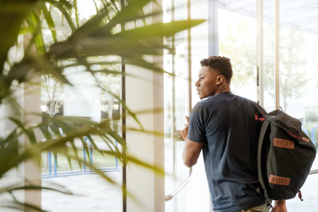 An image of a young male traveler with his back facing the camera as he pushes open a glass door to leave the airport after his first time flying. This image is used in the Students Fare student travel blog to share with first-time flyers, a guide to international flights for students!