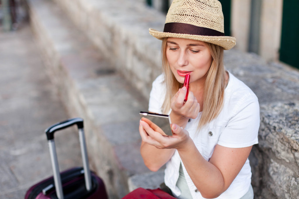 A blonde female traveler wearing a straw hat sits on a street curb with her suitcase, and applies red lipstick with a compact mirror. This image is used in the Students Fare student travel blog to share tips for packing luggage, "a guide to traveling with makeup and toiletries!"
