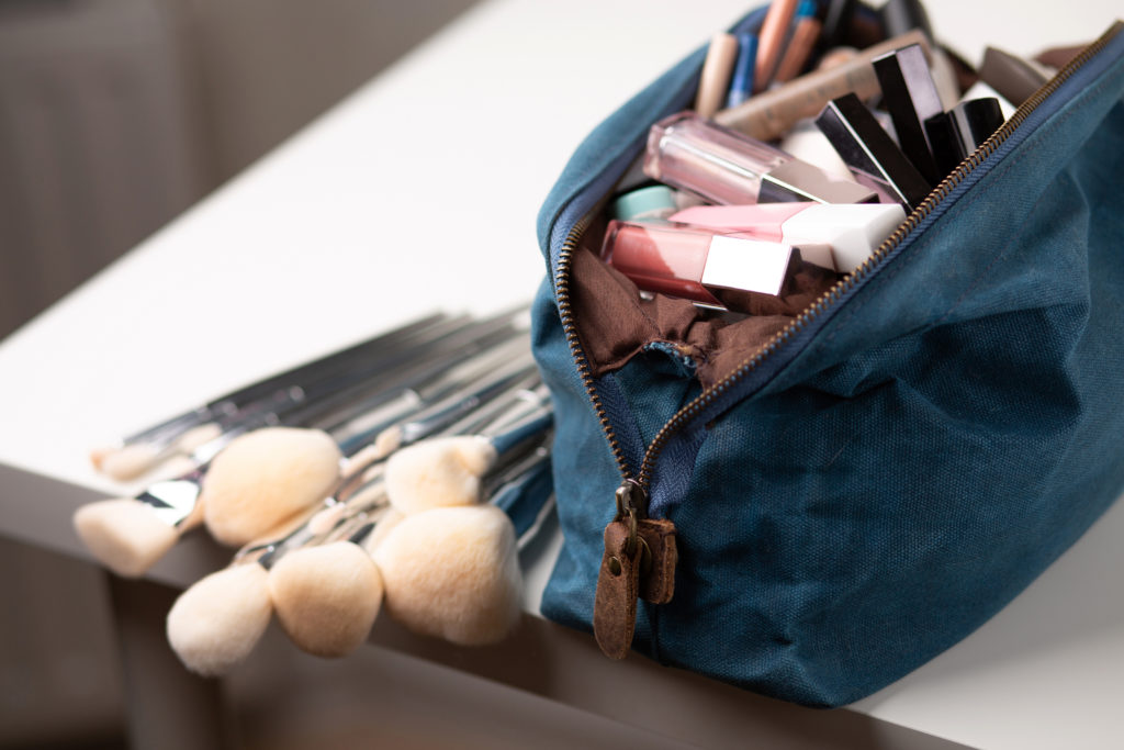 A blue makeup bag sits open on the table, revealing various products like lipstick, foundation, and blush, with brushes sitting next to it. This image is used in the Students Fare student travel blog to share tips for packing luggage, "a guide to traveling with makeup and toiletries!"