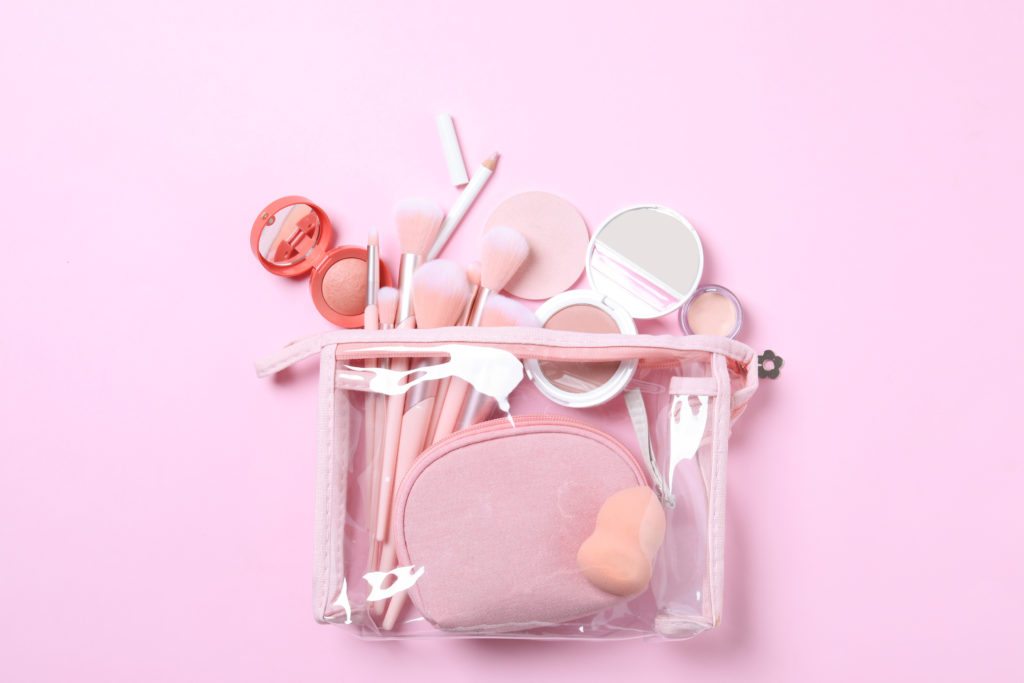 A clear travel bag sits on a flat pink surface with makeup brushes, compacts, and mirrors. This image is used in the Students Fare student travel blog to share tips for packing luggage, "a guide to traveling with makeup and toiletries!"
