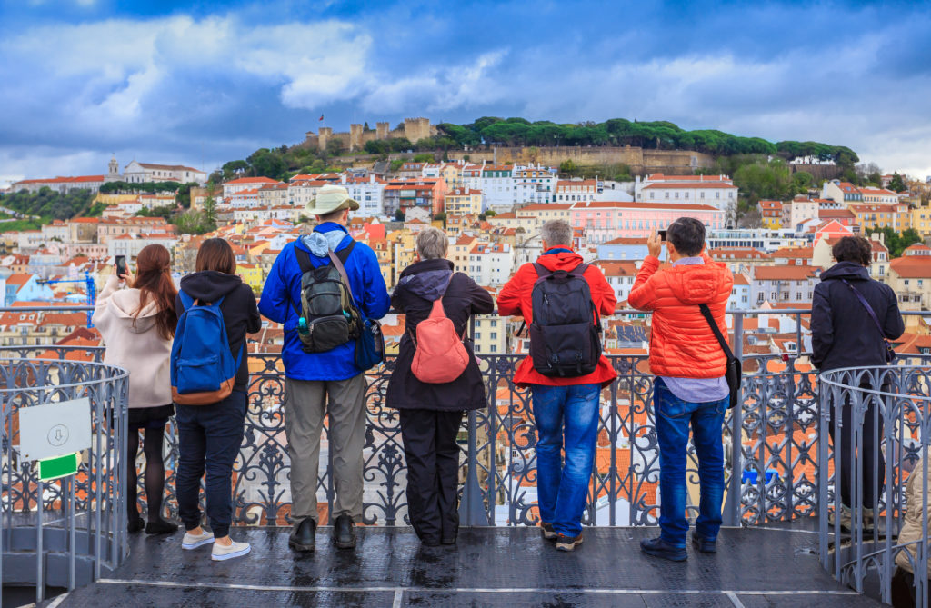 Cityscape of Lisbon, Portugal with pastel buildings climbing up a cliff and a large group of tourists taking photographs from a viewpoint. This image is featured in the travel blog by Students Fare, "Best Destinations for Group Vacations," which lists the top locations for youth and student groups to travel!