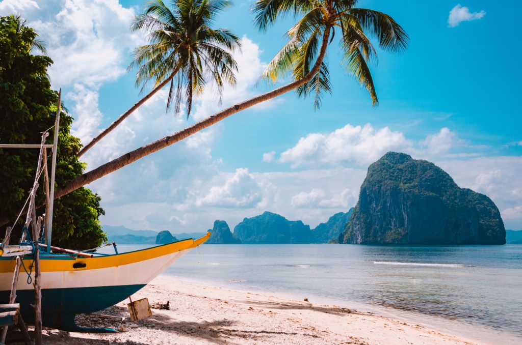 A beautiful landscape of Palawan, Philippines with a small yellow boat pulled into the sand with an island in the sunny distance. This image is featured in the travel blog by Students Fare, "Best Destinations for Group Vacations," which lists the top locations for youth and student groups to travel!