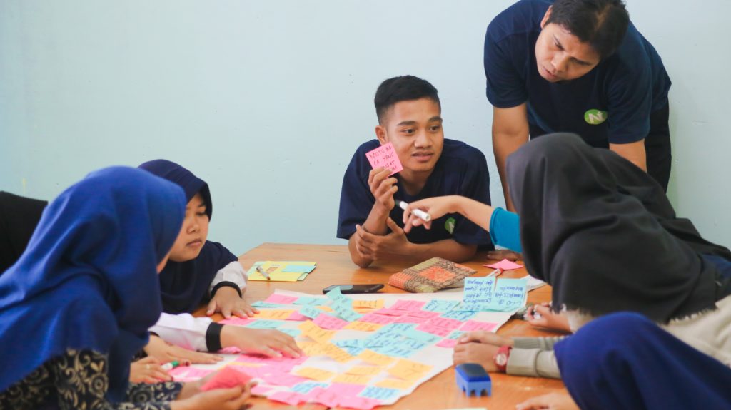 This image of a group of college students huddling over a table with sticky notes for planning is featured in the Students Fare travel blog, "Fundraising Ideas for Students," which describes different fundraisers for student and faculty to fund study abroad programs and school trips.