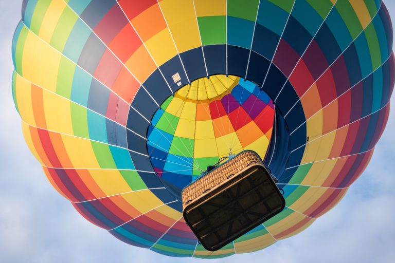 This closeup image of a hot air balloon with a rainbow pattern flying in the air is featured in the Students Fare travel and tour blog about tours, "Best Spots for Ballooning"