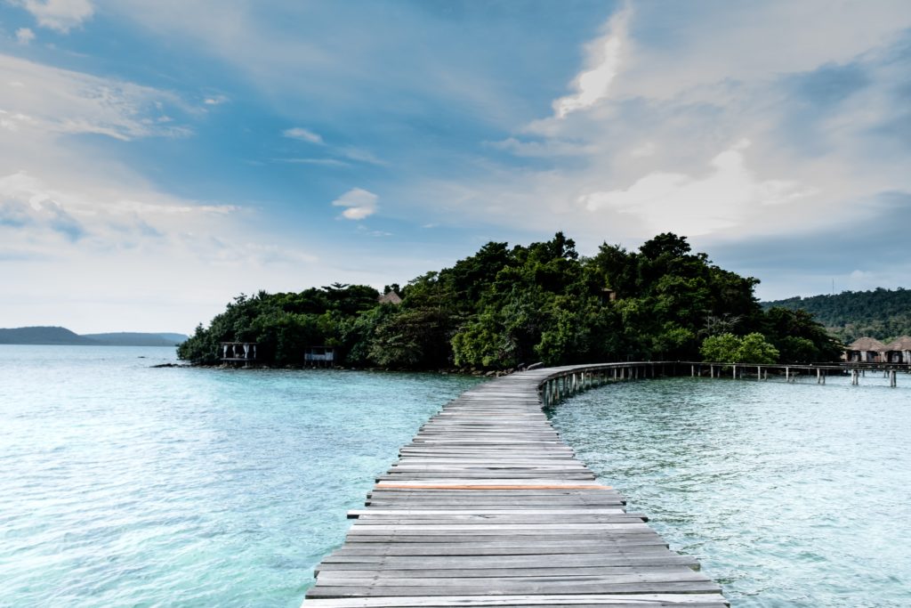 Featured in Affordable Vacation Destinations by Students Fare, this image shows a beautiful island in Cambodia with blue water and a bridge.