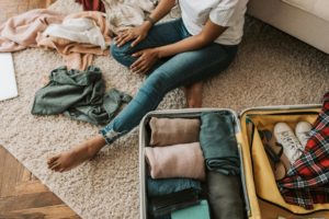Featured in Students Fare's blog article "Packing Tips for Student Travelers," this image shows a woman putting clothing into a yellow suitcase while sitting on the floor.